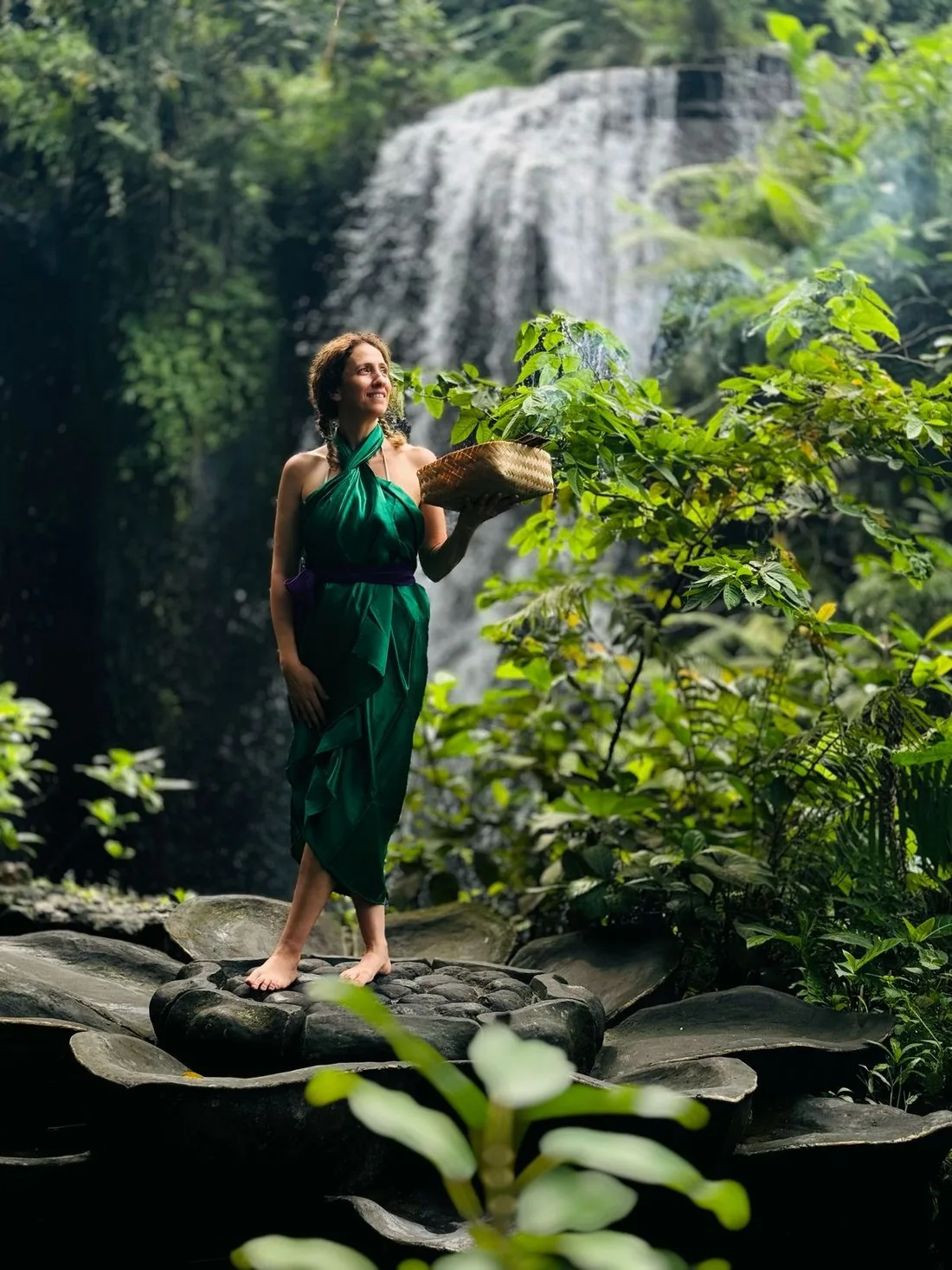 Tania standing barefoot on rocks in front of a waterfall, carrying a basket of greenery.